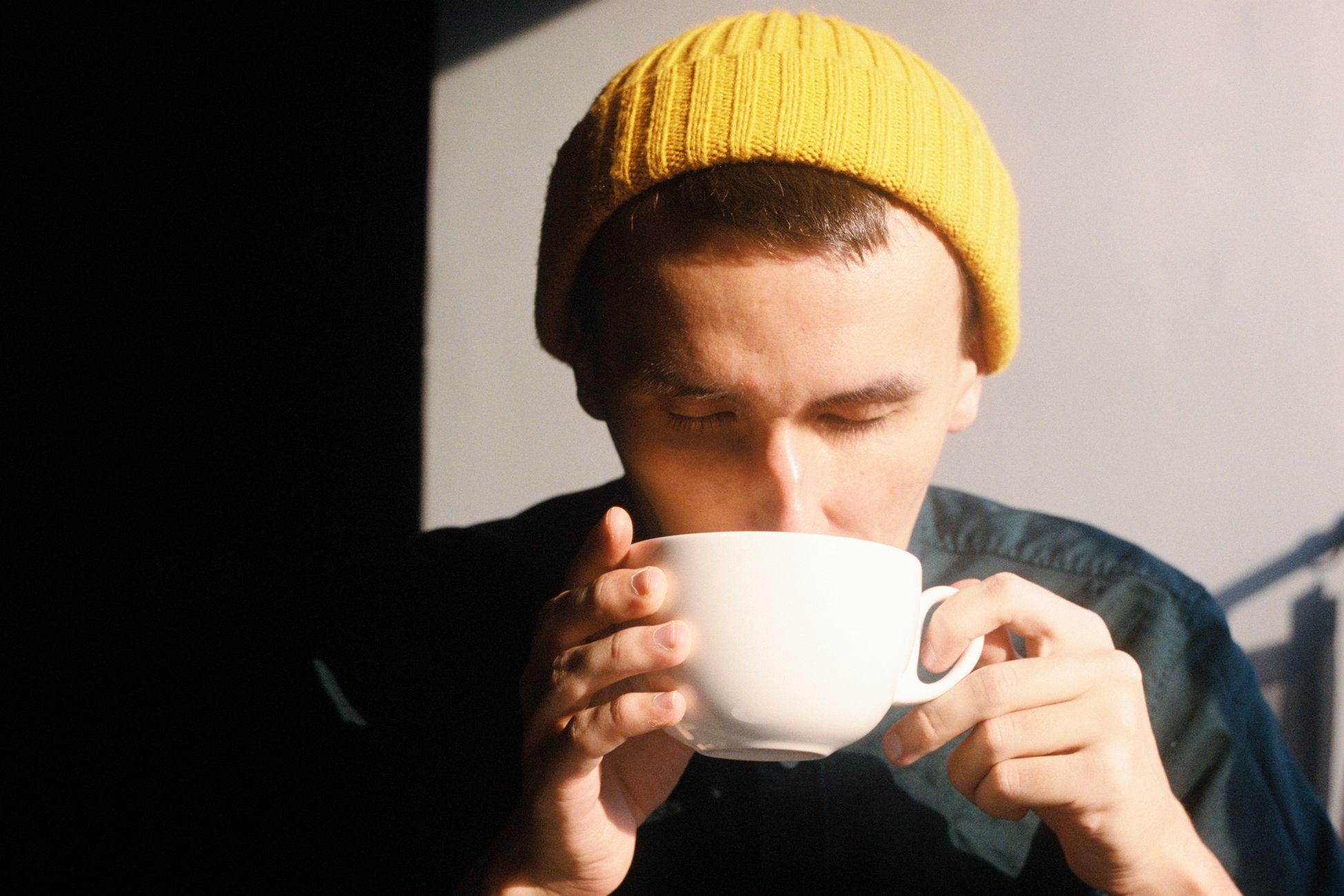 A young man enjoys a hot drink, wearing a yellow beanie in a sunlit room.