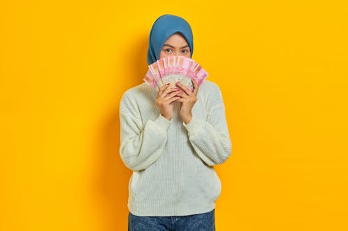 Free Beautiful Asian Muslim woman in white sweater covering face with cash money in Indonesian rupiah banknotes isolated over yellow background. ​People religious lifestyle concept Stock Photo