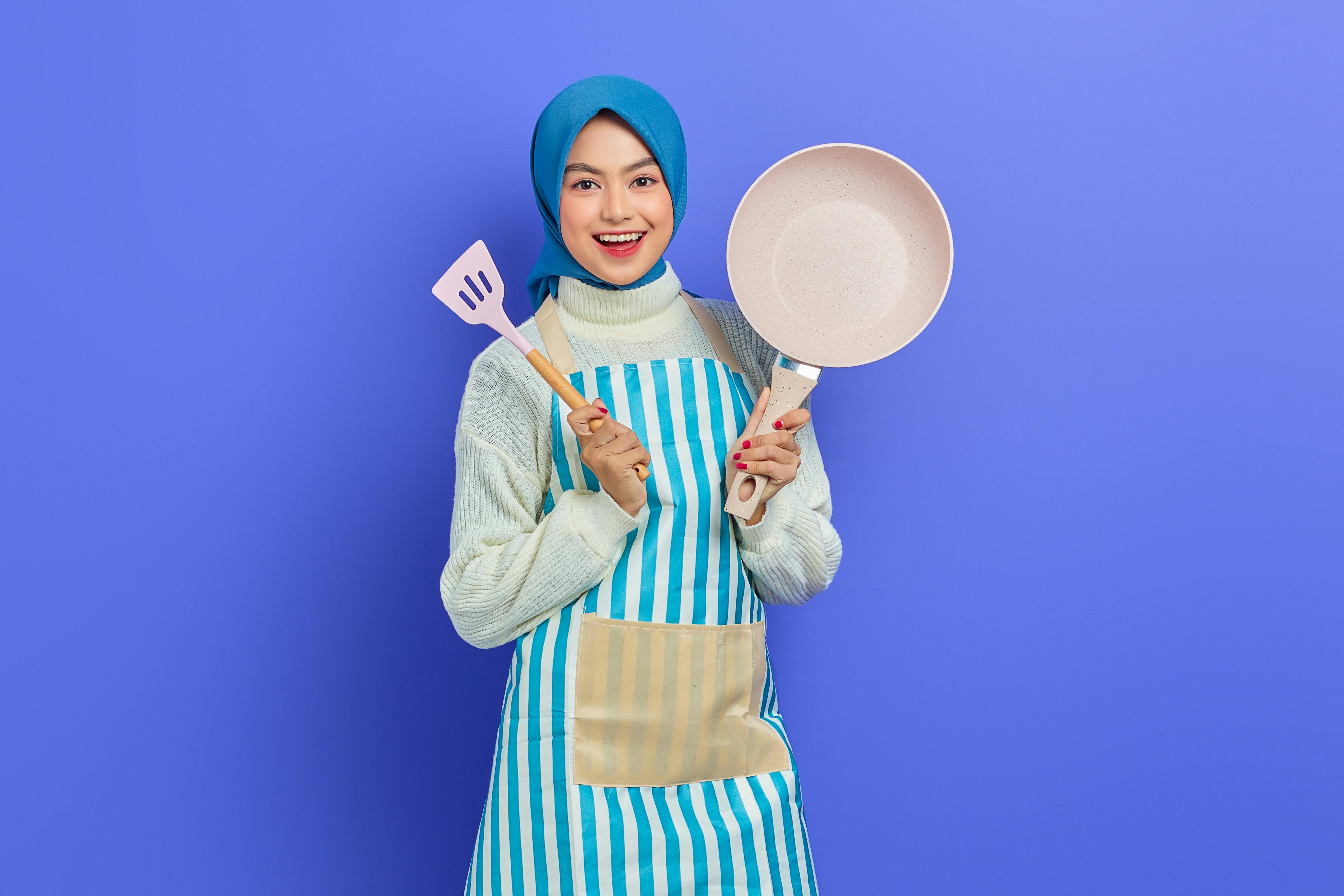 a young woman wearing blue hijab and apron holding a frying pan and slotted turner