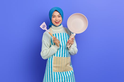 Cheerful woman in hijab with pan and spatula, ready for cooking. Studio shot against blue background.