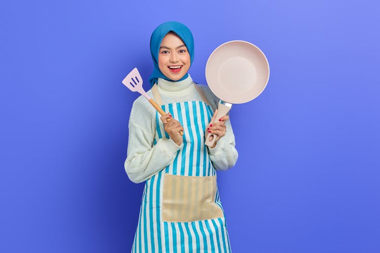 A Young Woman Wearing Blue Hijab And Apron Holding A Frying Pan And Slotted Turner