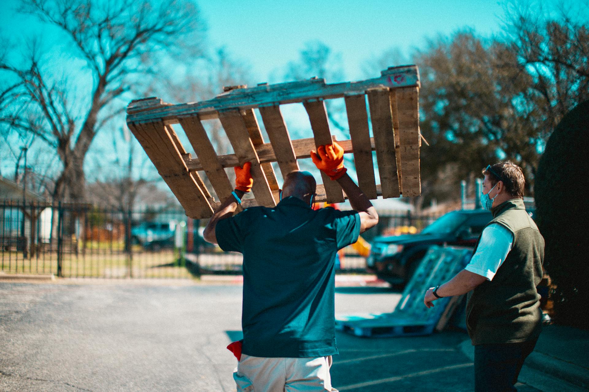 A man wearing gloves carries a wooden pallet in an urban area, showcasing manual labor.