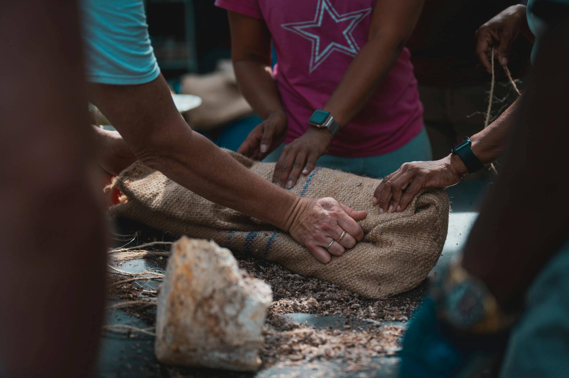 Hands working together to secure a burlap sack on a table, showcasing teamwork.
