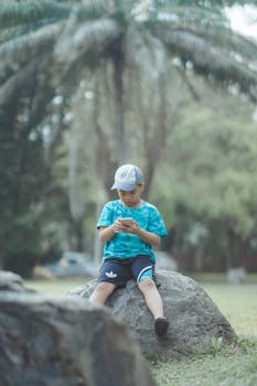 A child in a green shirt is sitting on a rock in a park using a smartphone, enjoying a leisurely outdoor day.
