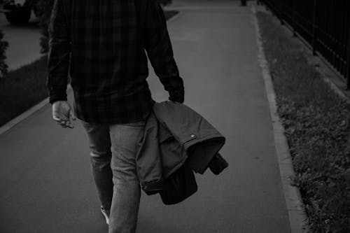 Grayscale Photo of a Person Holding a Jacket while Walking on the Street