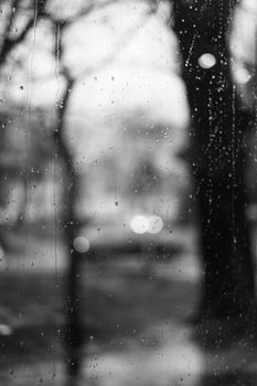 Atmospheric black and white photo of rain on a window with blurred trees beyond.