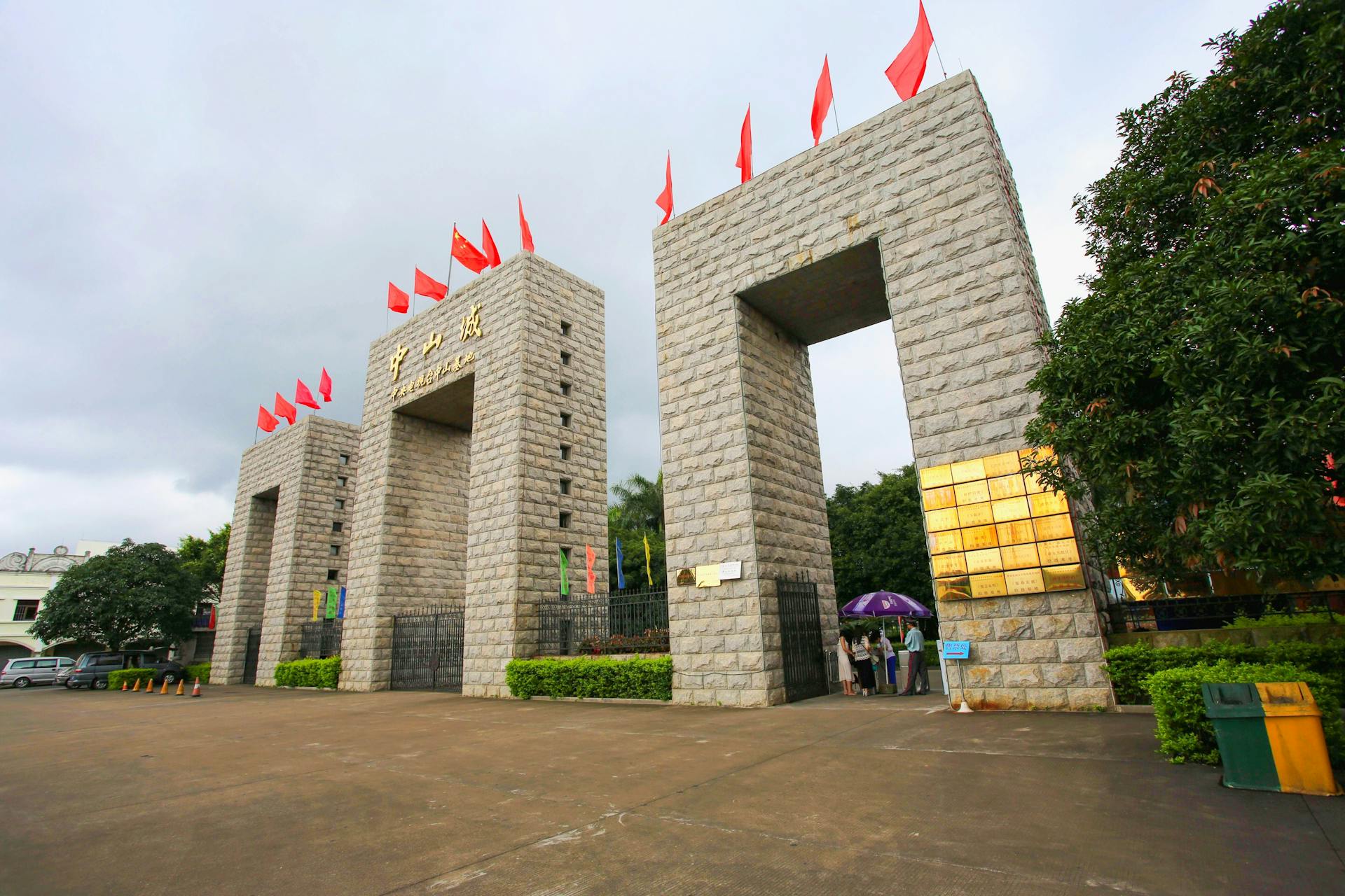 Entrance gate with red flags and Chinese script under cloudy sky, showcasing cultural architecture.