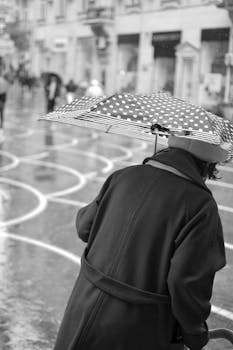Black and white photo of an elderly person with a polka dot umbrella walking on a rainy city street.
