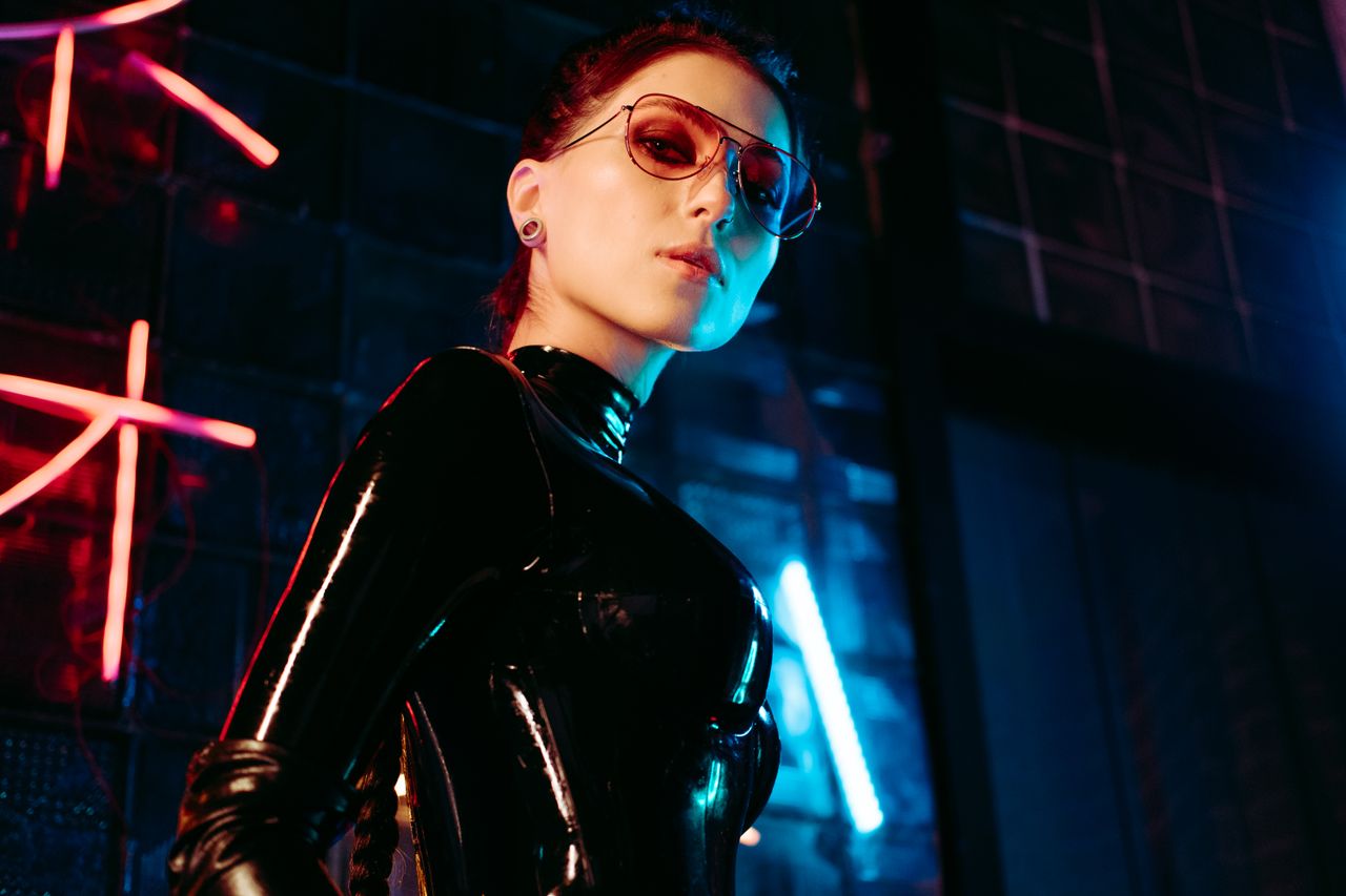 woman in glossy leather outfit with sunglasses posing against neon lit