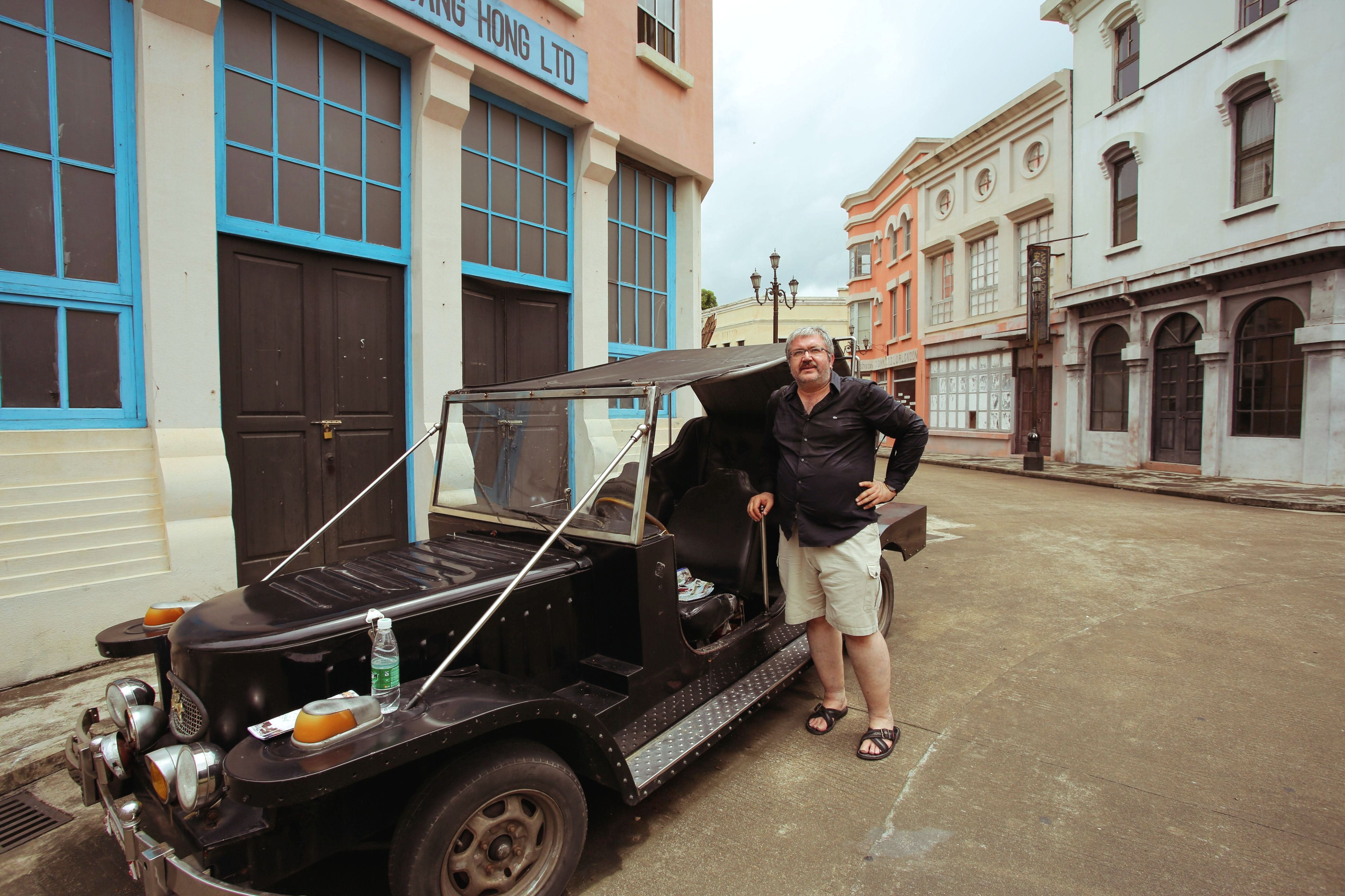 A man poses with a vintage car on a street lined with colonial-style buildings.