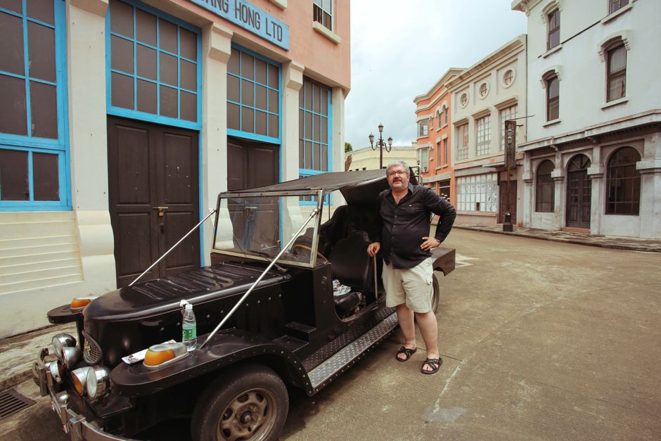 A man poses with a vintage car on a street lined with colonial-style buildings.