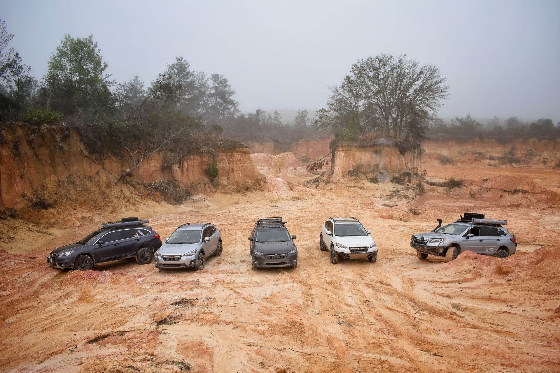 Aerial view of five off-road vehicles on a foggy, desert-like terrain surrounded by trees.