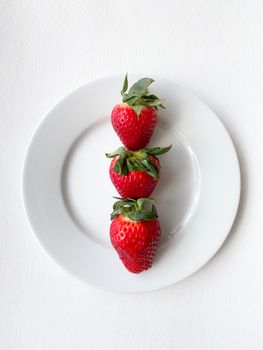 Three ripe strawberries neatly arranged on a white plate against a bright background.