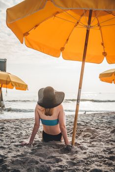 Woman in sunhat relaxing on a sandy beach under vibrant umbrellas by the sea.