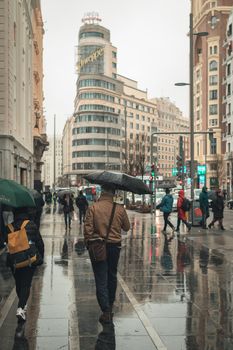 People with umbrellas walking on a wet street in Madrid, featuring iconic Schweppes building.