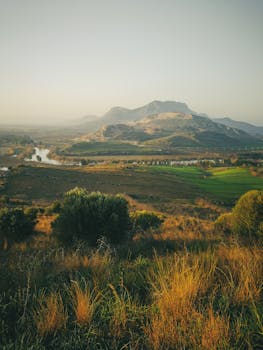 A beautiful countryside view featuring green fields, a calm river, and majestic mountains in the distance, captured at sunset.