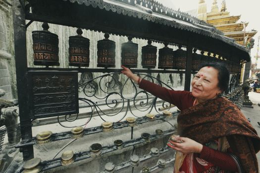 Elderly woman engaging with prayer wheels in a traditional Asian temple, expressing cultural devotion.