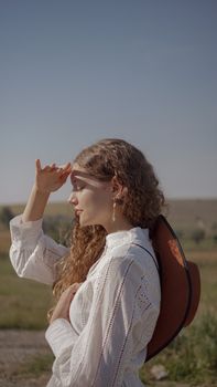 Woman with curly hair shielding eyes from sun in open field, wearing white top.