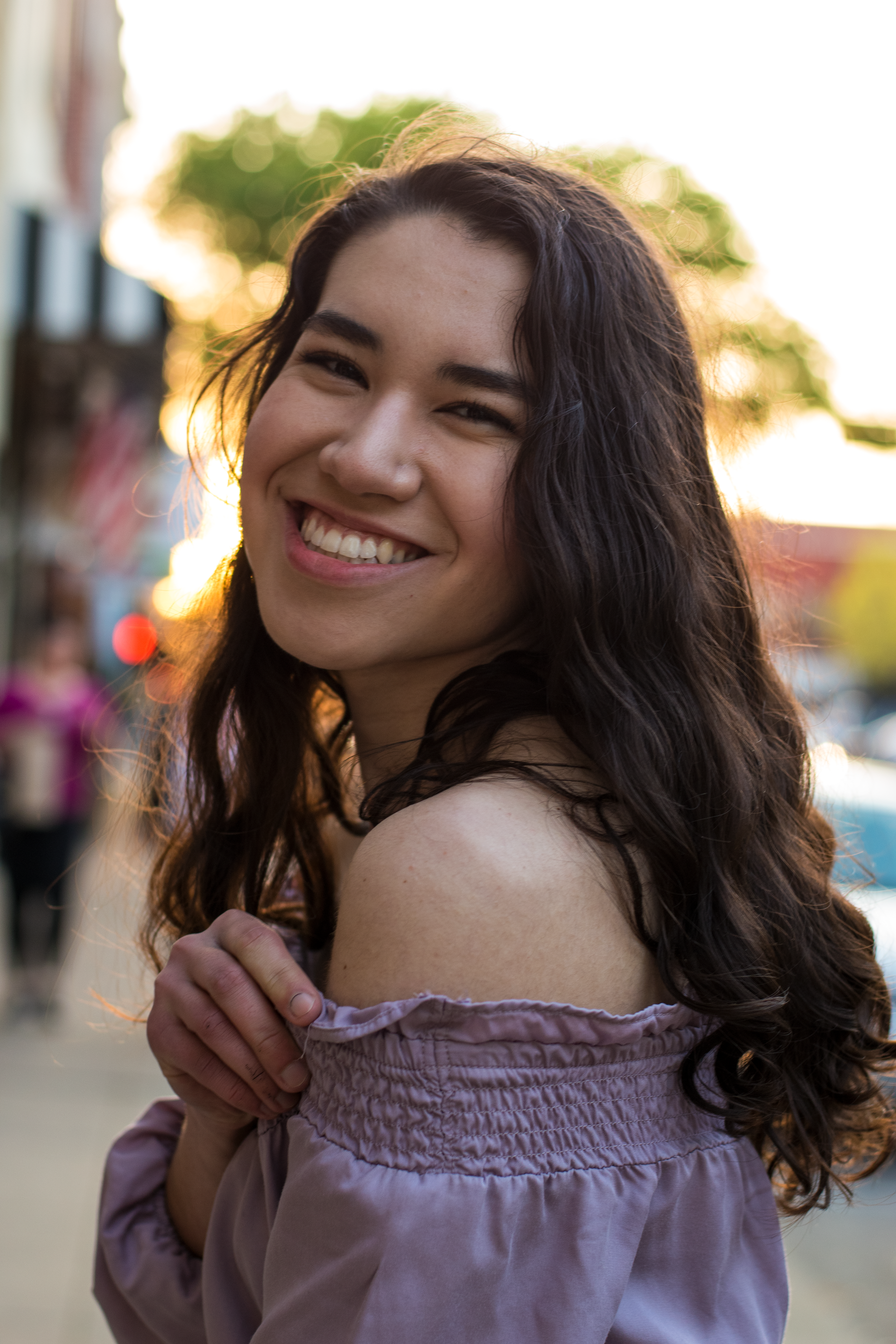 Backlit portrait of a smiling woman wearing a purple off-shoulder top on a vibrant city street during sunset.