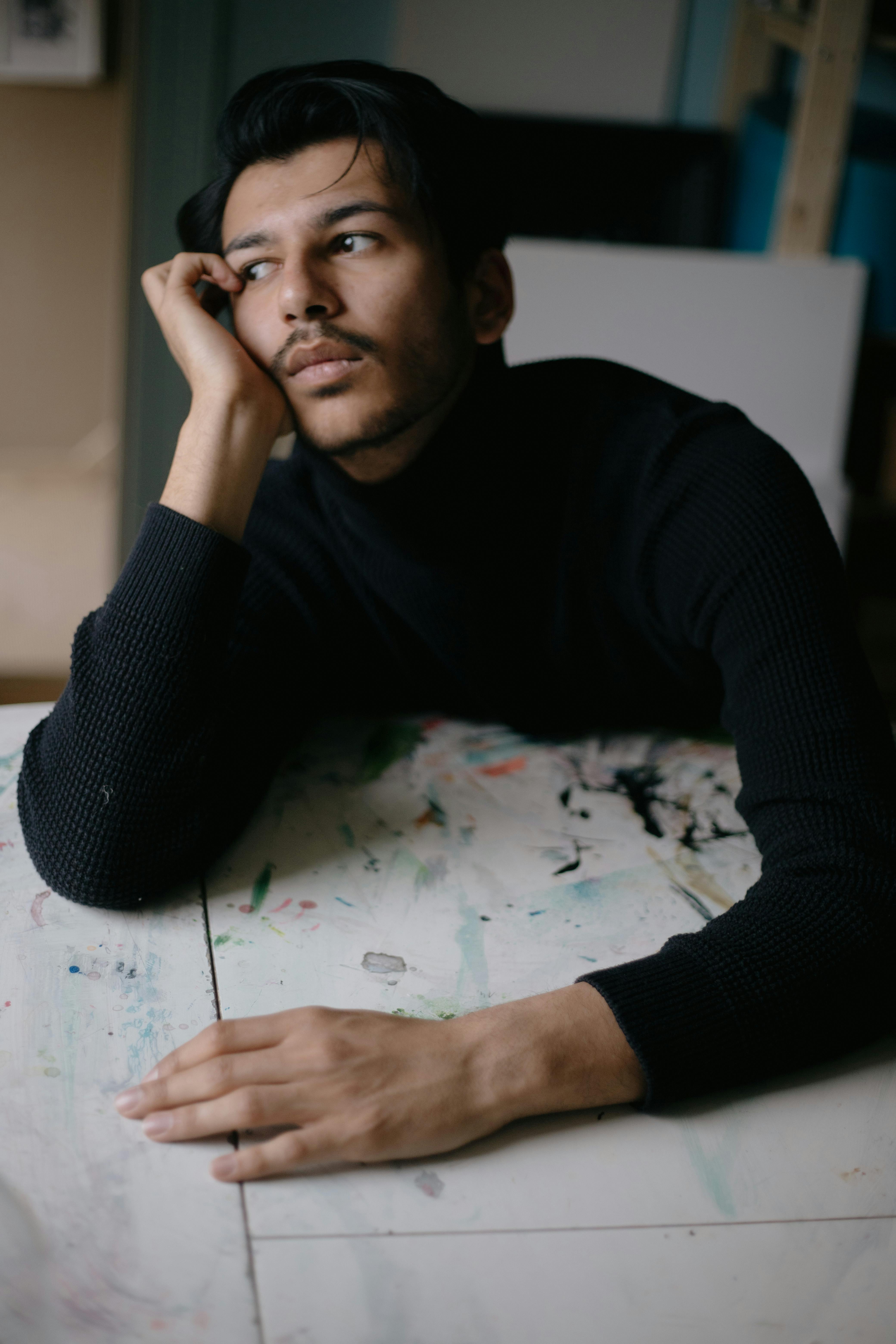 A young man in a black sweater rests his chin on his hand, looking thoughtful.