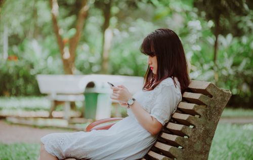 Free A young woman in a white dress sitting on a park bench, using a smartphone. Stock Photo