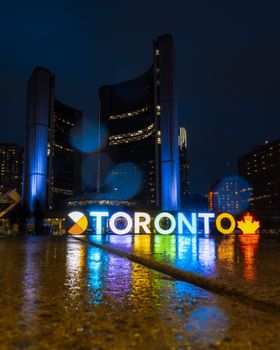 Illuminated Toronto sign and City Hall at night with rain reflections.