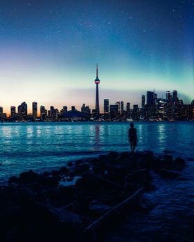 Silhouette of a person by the water with Toronto's illuminated skyline and CN Tower at twilight.