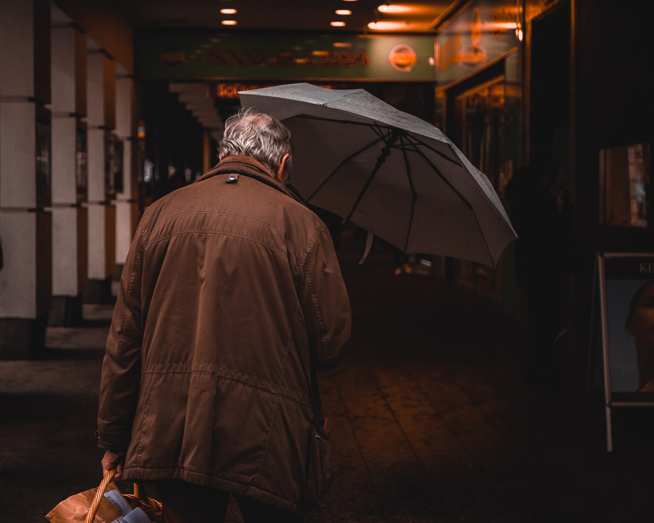 A senior man in a brown jacket walks down a Vienna street holding an umbrella on a rainy day, illustrating Travel Insurance for Seniors — 1-800-MEDIGAP, America's Trusted Toll-Free Number.
