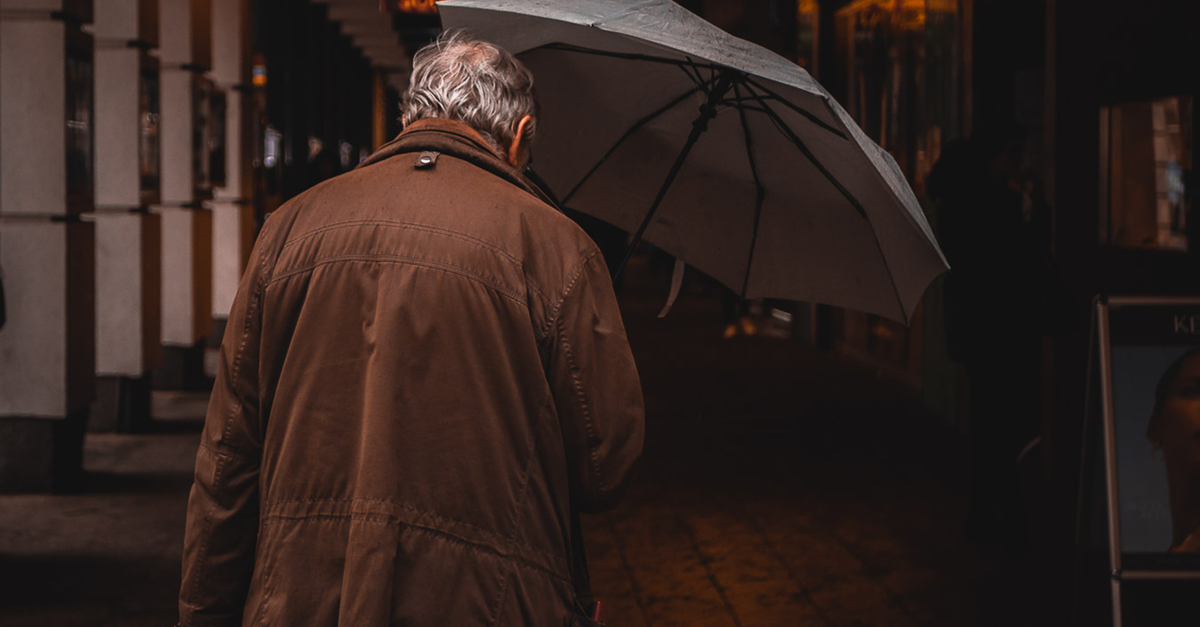 A senior man in a brown jacket walks down a Vienna street holding an umbrella on a rainy day, illustrating Travel Insurance for Seniors โ 1-800-MEDIGAP, America's Trusted Toll-Free Number.