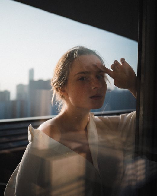 Artistic portrait of a woman looking through a window with cityscape background, creating an introspective mood.