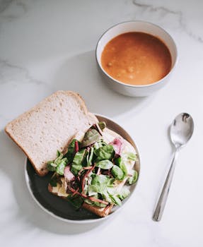 Close-up of a healthy greens sandwich paired with tomato soup on a white table setting.