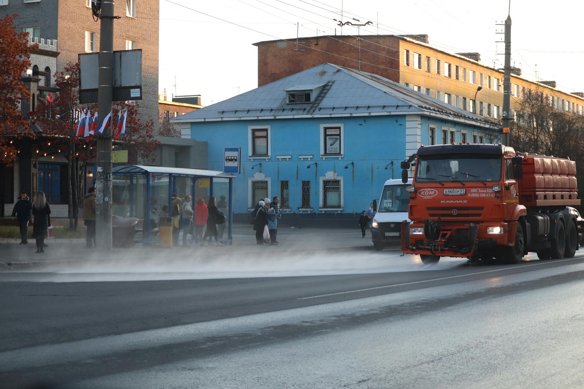 An orange street cleaning truck sprays water on a city road near a bus stop with people waiting.