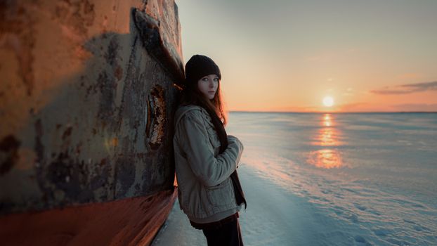 A woman in a beanie leans against a rusted structure on a frozen beach during sunset.