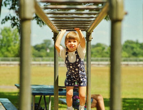 A Girl in White Shirt and Floral Jumper Playing on a Monkey Bars