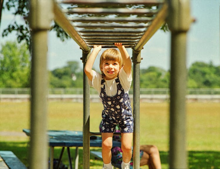 A Girl In White Shirt And Floral Jumper Playing On A Monkey Bars