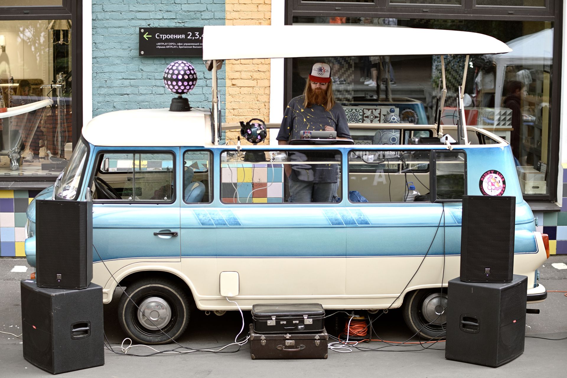 DJ playing music from a vintage van at a street performance with speakers and disco balls.