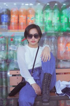Fashionable woman in sunglasses sitting on a bench with colorful drink bottles background.