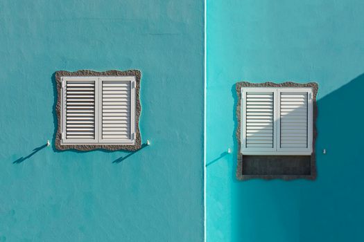 Minimalist blue wall exterior featuring two white shuttered windows casting shadows.