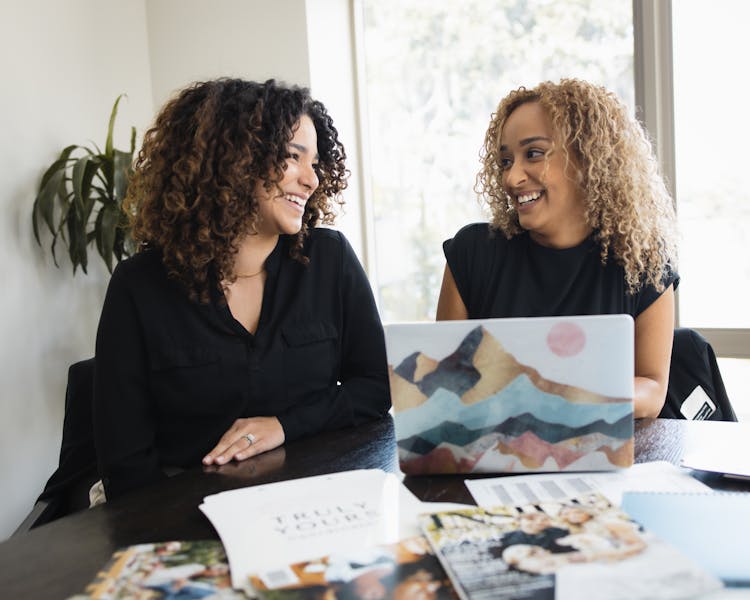 Women Sitting Behind A Table Looking At Each Other And Laughing