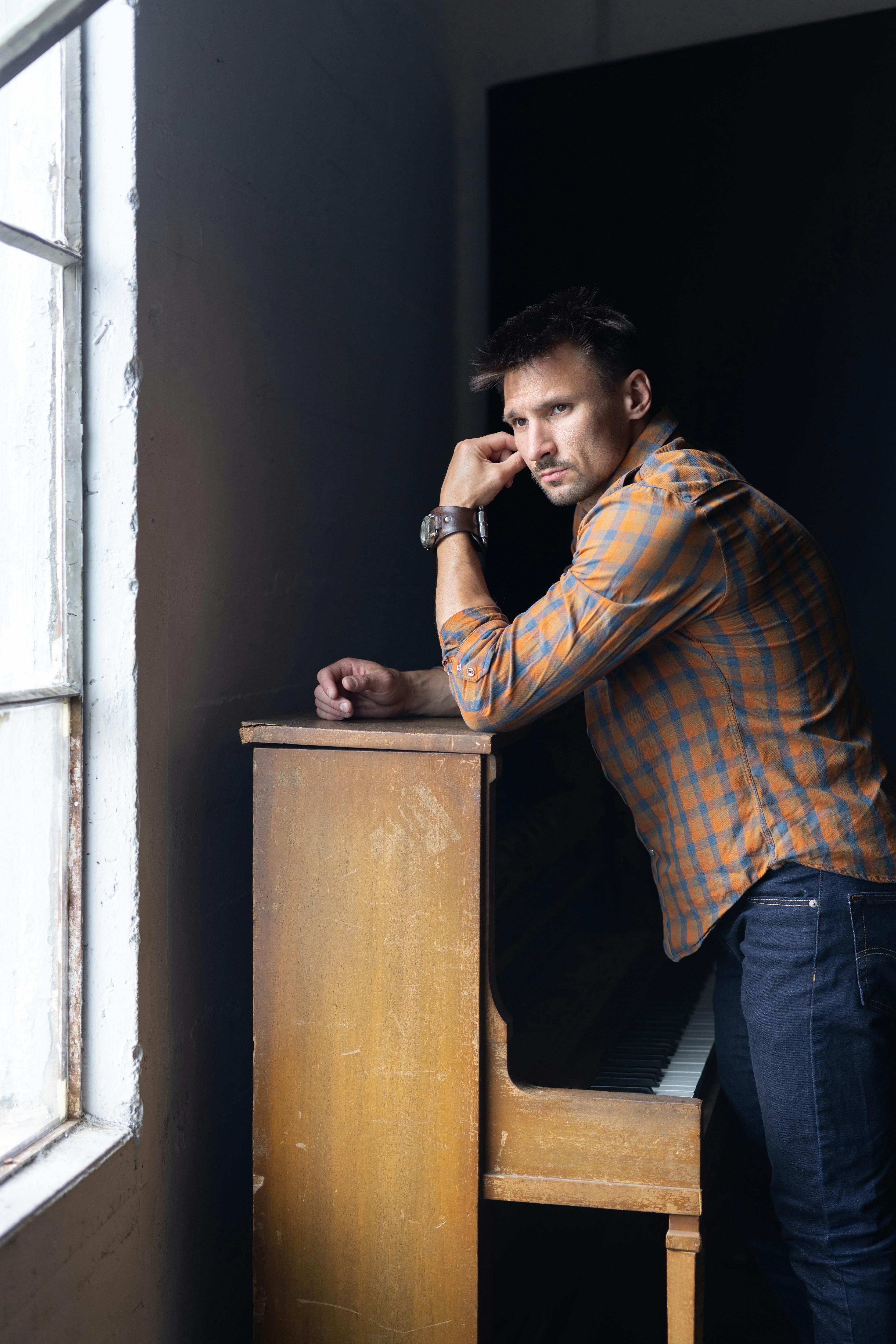 A man in a plaid shirt stands indoors, leaning on a piano, gazing outside with a thoughtful expression.
