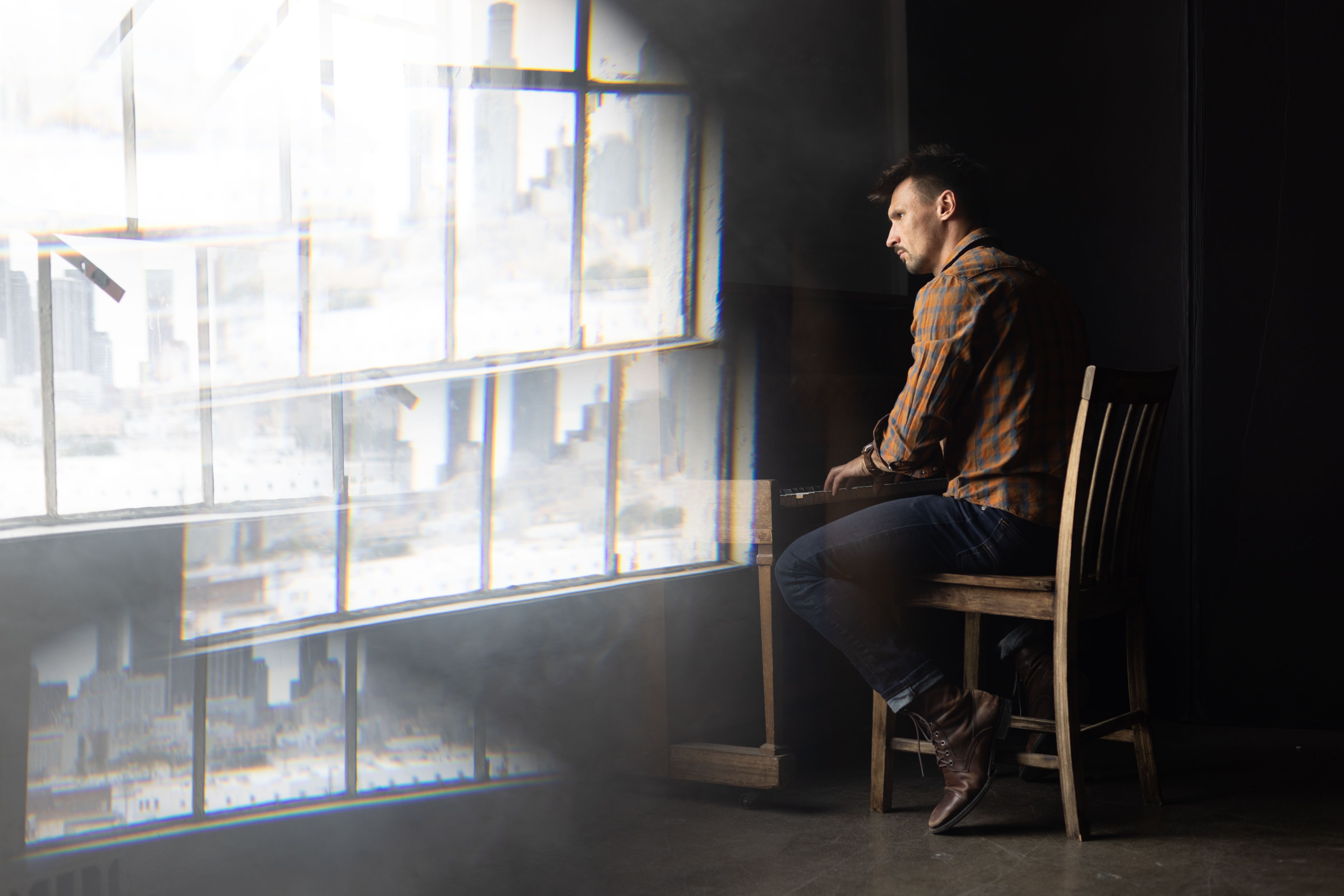 A man in a plaid shirt sits thoughtfully at a piano by a large window indoors.
