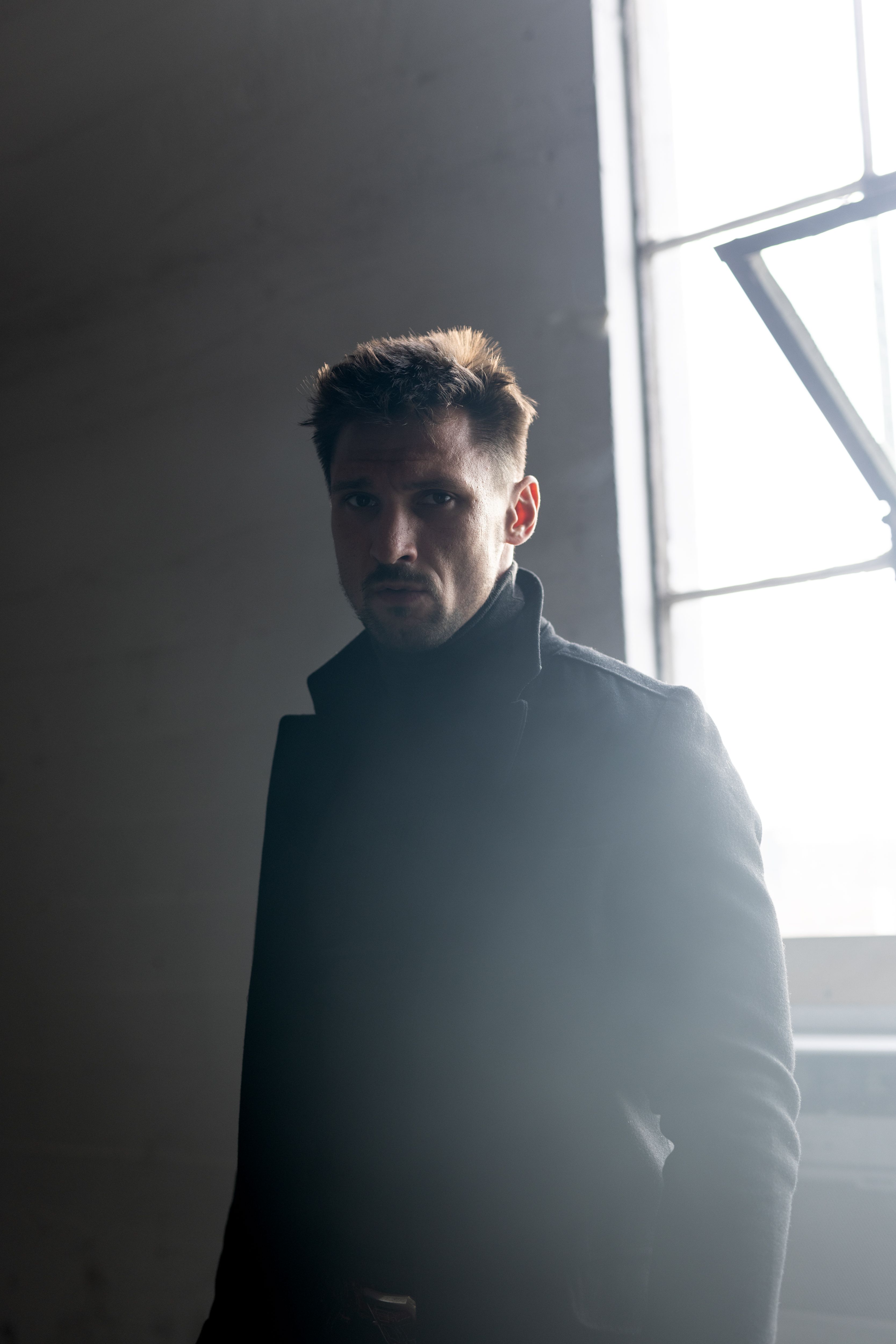 A contemplative man stands by a large window, casting dramatic shadows indoors.