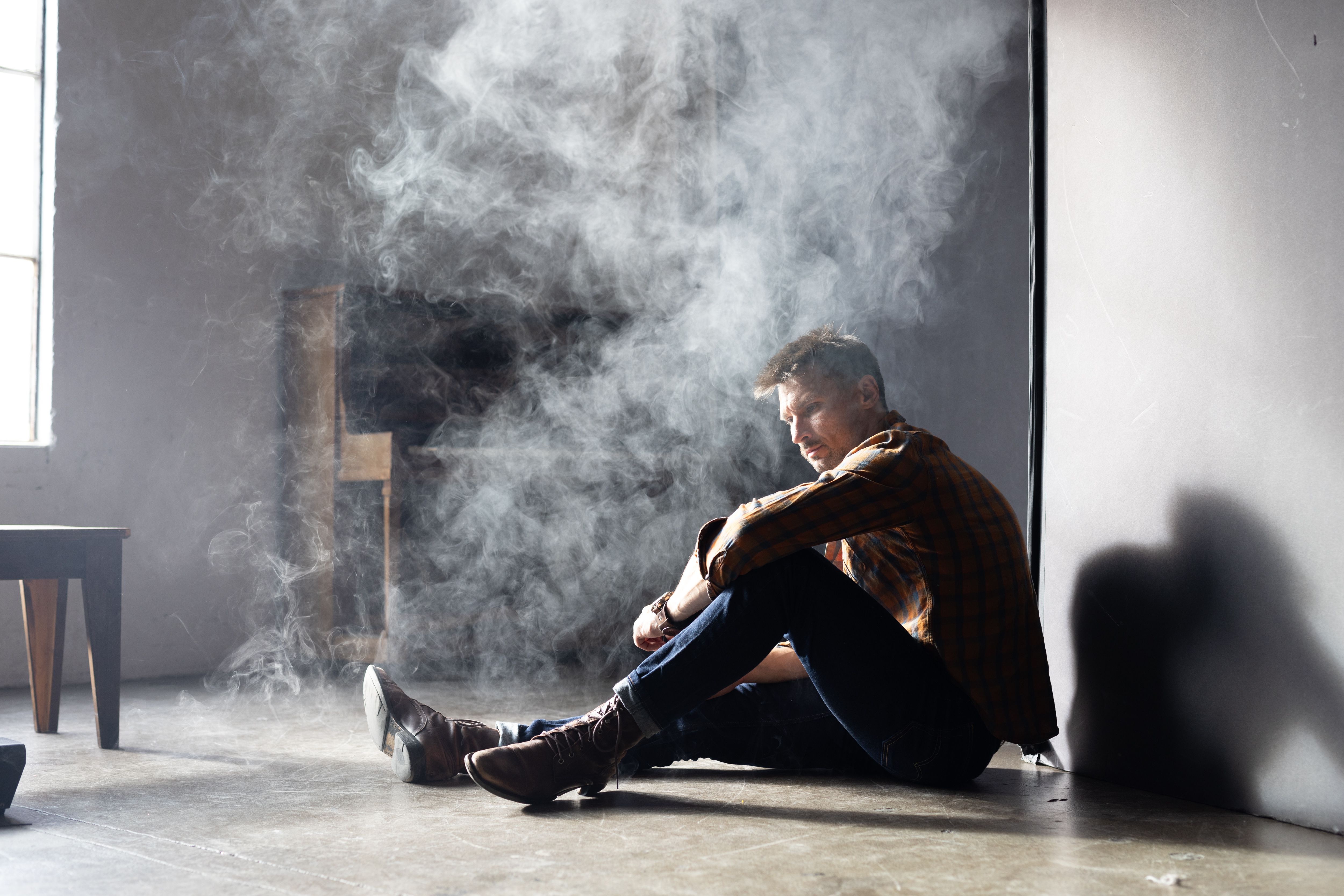A man in boots and denim sits pensively in a smoke-filled room with soft natural light.