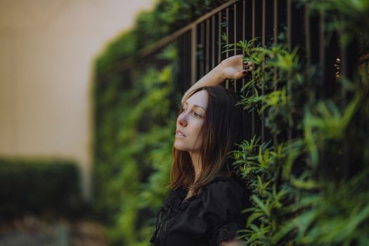 Woman with long hair posing by a lush green foliage fence, creating a serene atmosphere.