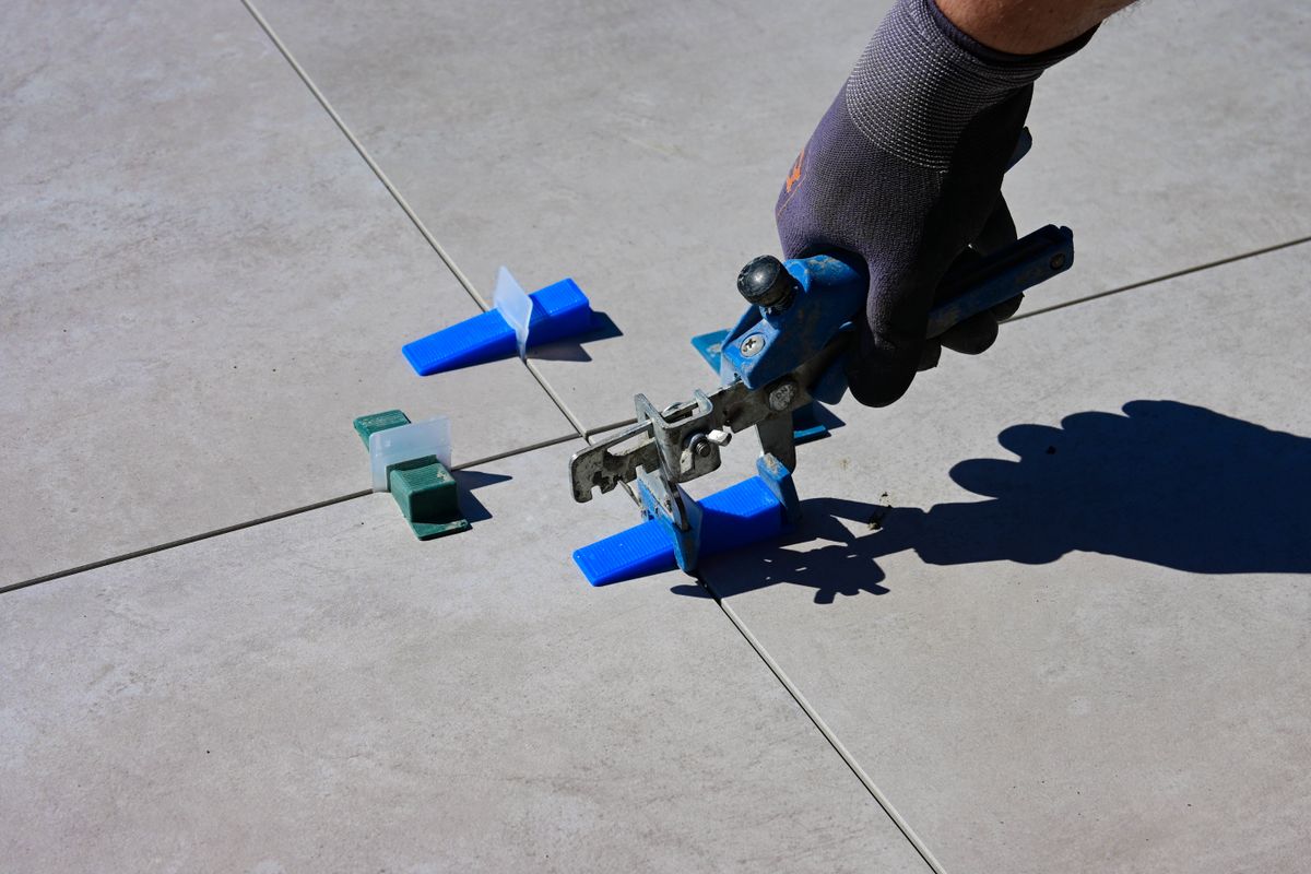Gloved worker using a blue tile-levelling tool on large outdoor floor tiles, with clips and wedges nearby.