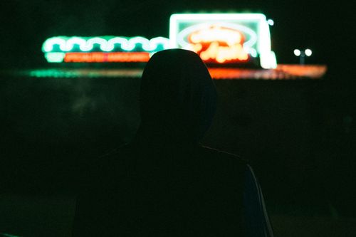 Free A mysterious figure in a hoodie standing at night against a glowing neon sign. Stock Photo