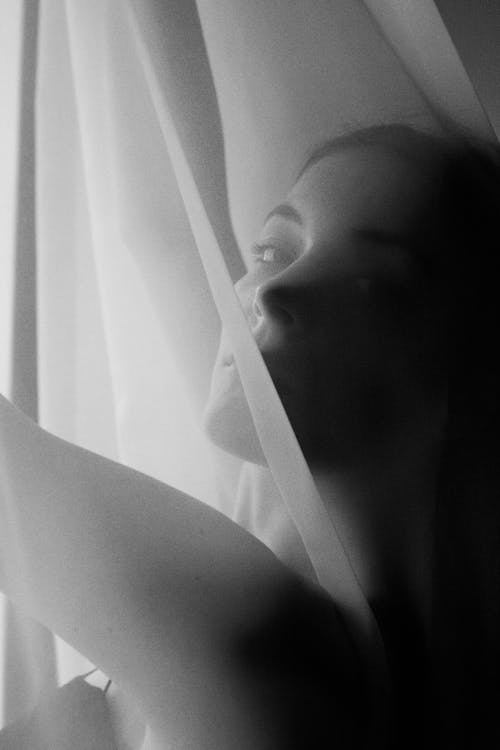 Grayscale Photo of a Woman Behind a Curtain