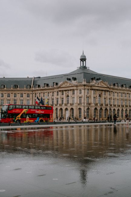 Fotomural Edificio neoclásico reflejando en el agua con un autobús turístico rojo en una ciudad.