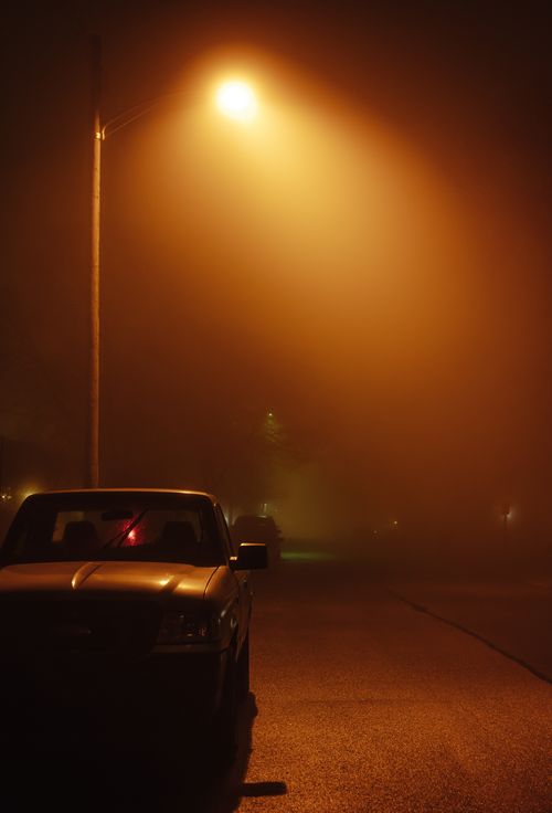 Free Eerie city street at night illuminated by a glowing street lamp, casting shadows on a parked car. Stock Photo