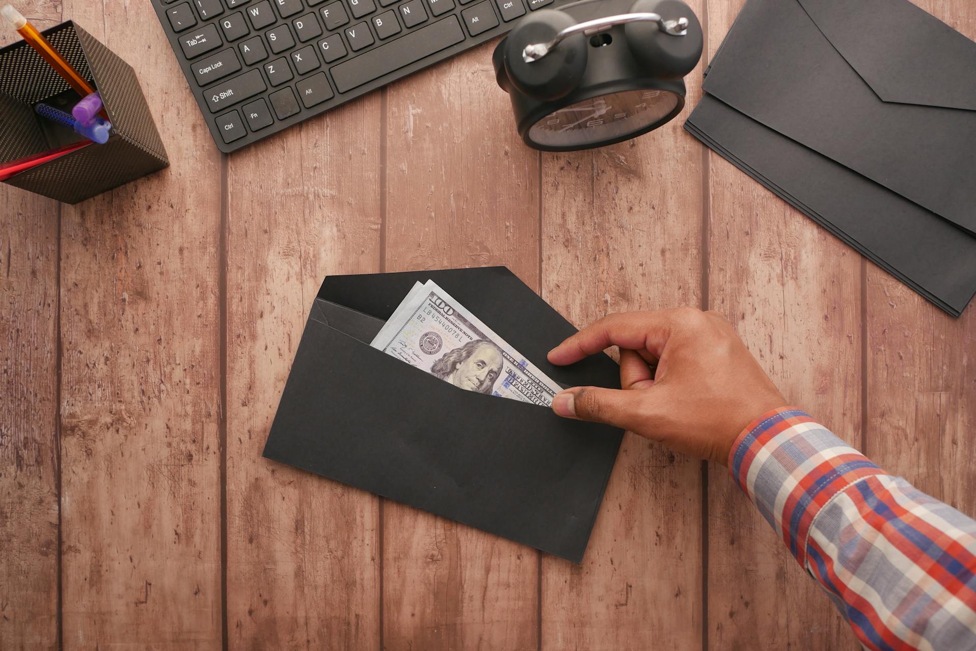 A hand placing a 100 dollar bill in a black envelope on a wooden desk with stationery, keyboard, and alarm clock.